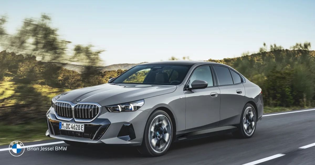 A silver BMW 5 Series sedan driving on a highway with blurred trees and hills behind.
