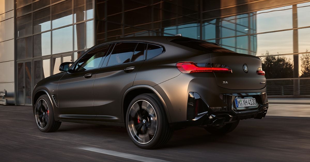 Matte black BMW X4 M Competition drives past modern architecture, highlighting sleek coupe styling and aggressive performance stance