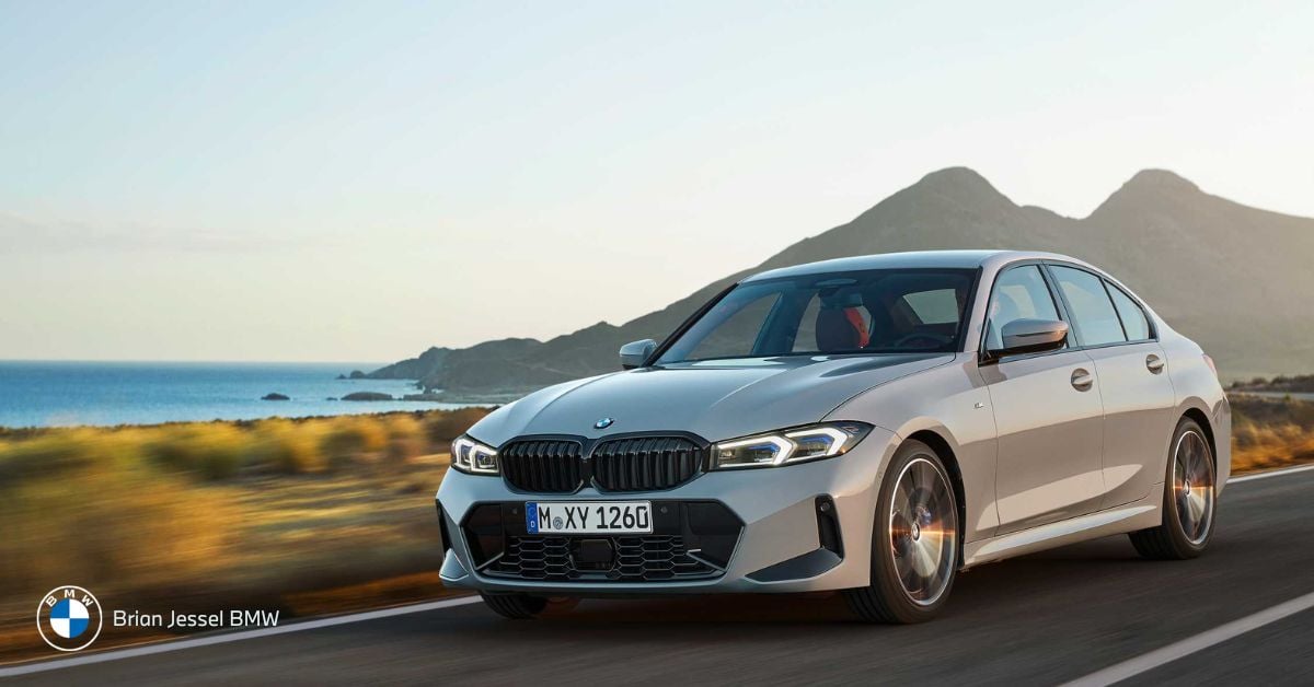 A silver BMW sedan driving on a coastal highway with mountains and ocean in the background.