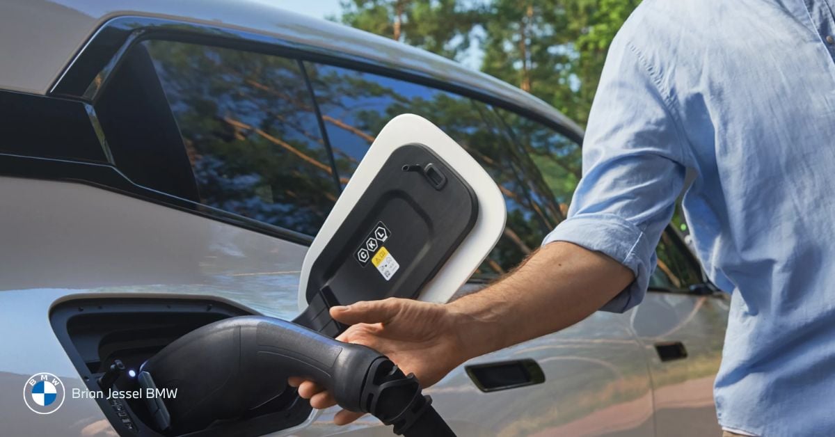 Person plugging electric vehicle charging cable into BMW parked outdoors near trees during daytime summer
