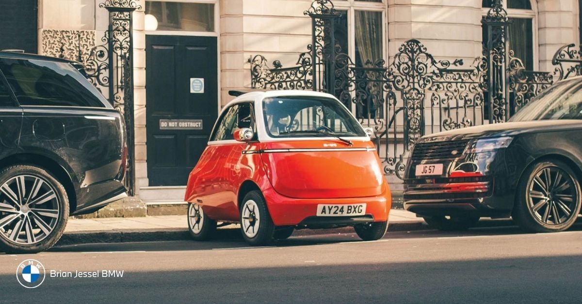 Red BMW Isetta parked between two large modern black SUVs on a city street, showing dramatic size contrast.