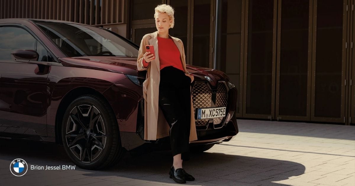 Blonde woman in red top leaning on maroon BMW SUV, checking remote software update on smartphone outdoors.