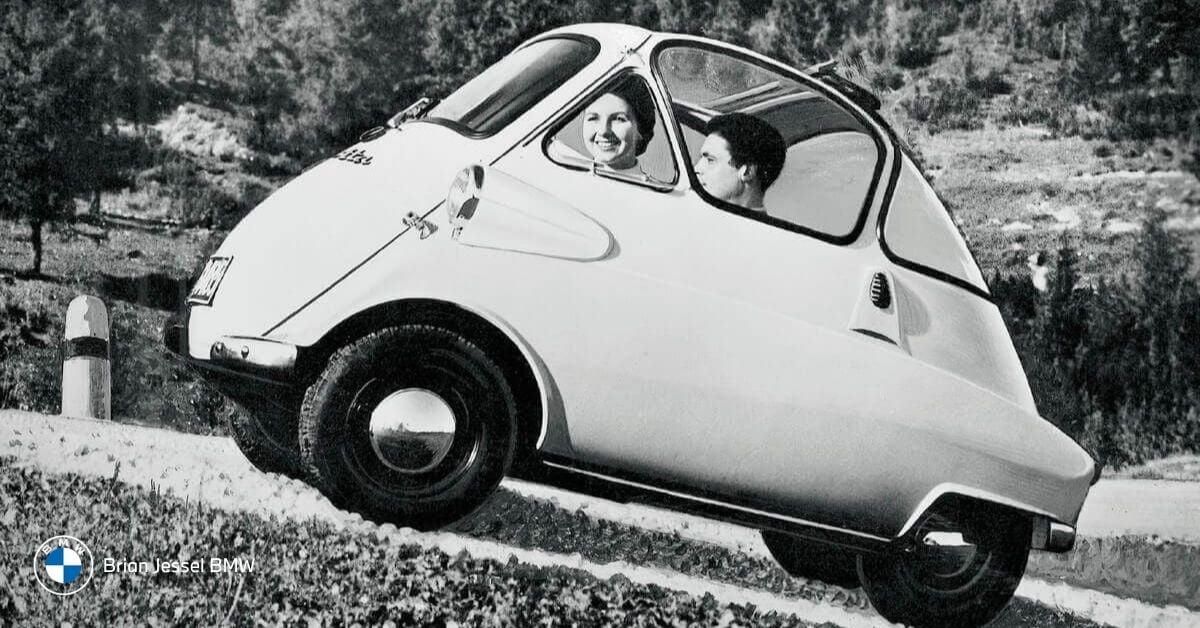 Classic white BMW Isetta bubble car with a smiling couple driving on a scenic countryside road.