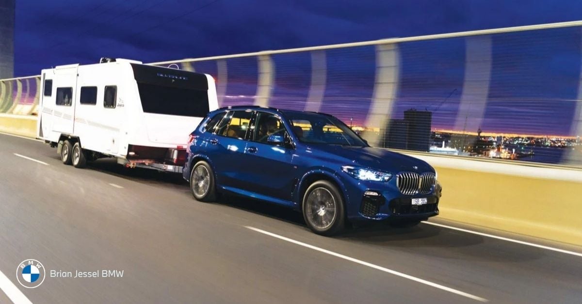 Blue BMW X5 SUV towing a large white caravan trailer on a highway at dusk