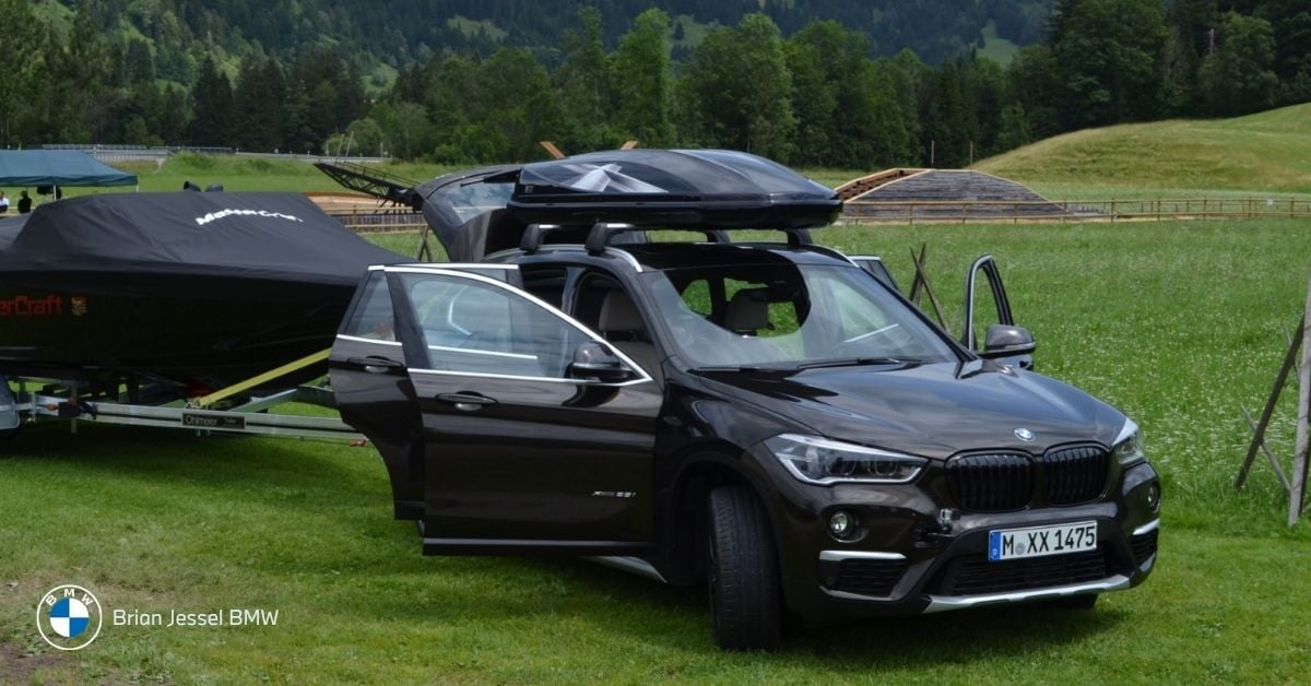 BMW X1 SUV with open doors and roof box parked on grass towing a covered boat trailer