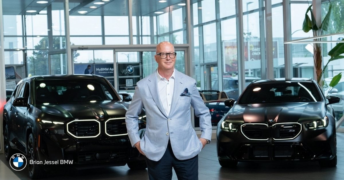 Smiling man in light blue blazer standing between two black BMW iX3 electric SUVs in Brian Jessel BMW showroom.