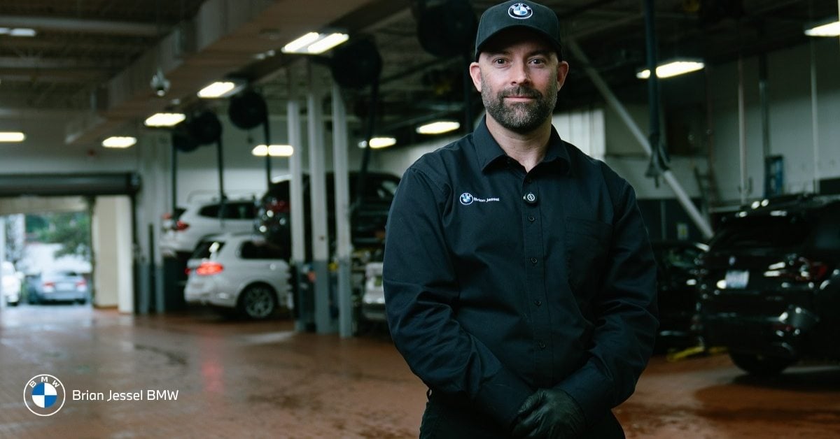 Smiling BMW technician in black uniform and cap standing confidently in the Brian Jessel BMW Vancouver service garage, with vehicles and service bays visible in the background.