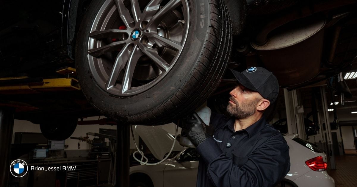 BMW technician inspecting wheel and suspension components beneath raised vehicle during professional dealership maintenance service appointment.
