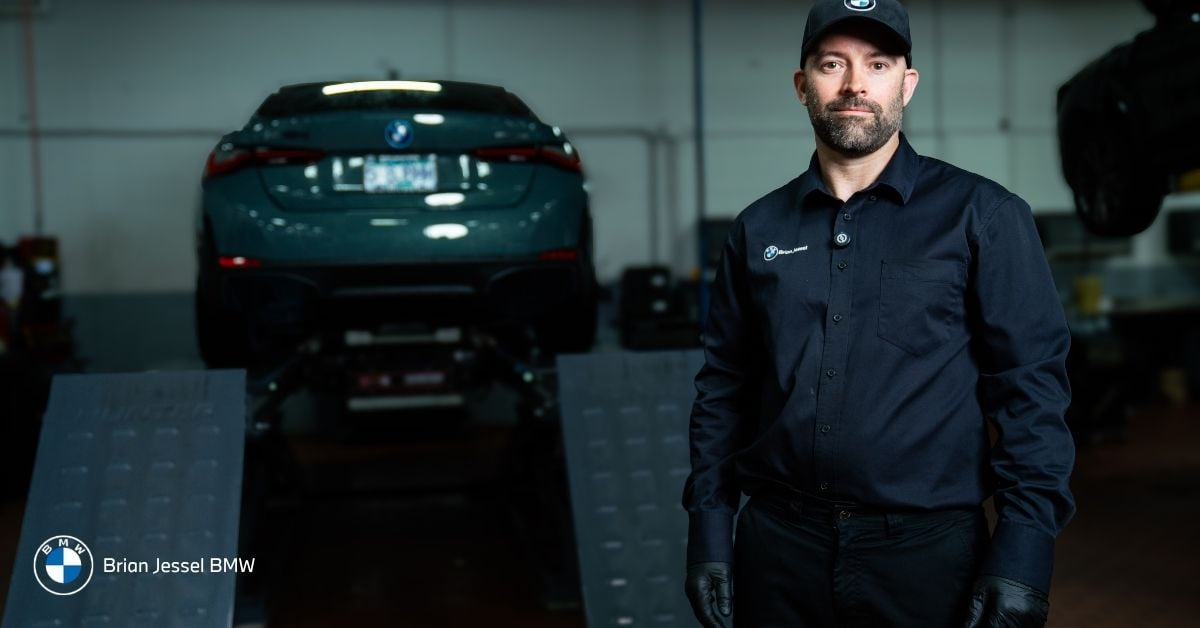 BMW technician in uniform and cap stands in service bay with green BMW elevated on lift.