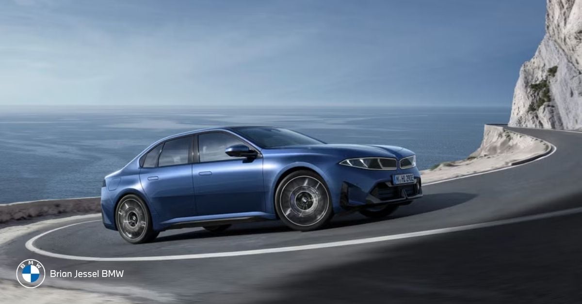 Blue BMW Neue Klasse sedan i3 50 xDrive drives along scenic coastal cliff road overlooking the ocean under clear skies.