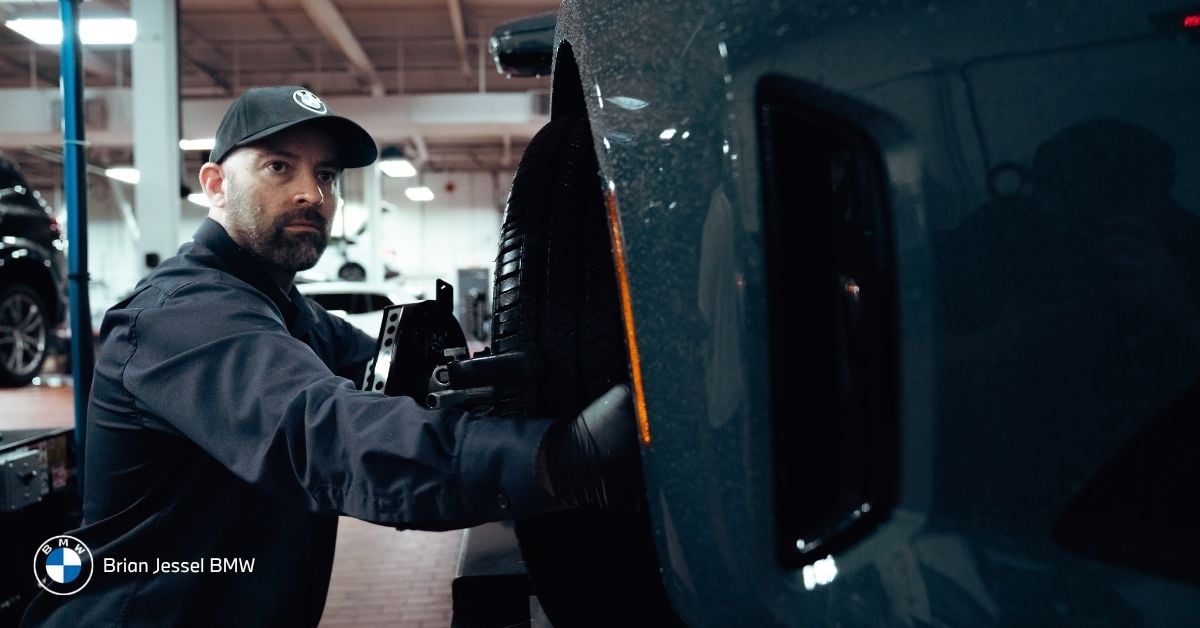 BMW technician inspects rear tire of dark vehicle during scheduled maintenance service in dealership workshop bay.