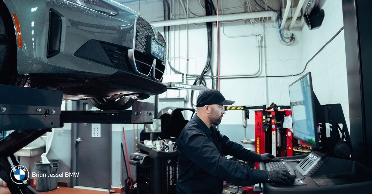 Technician works at computer beside elevated gray BMW in service bay during scheduled maintenance.