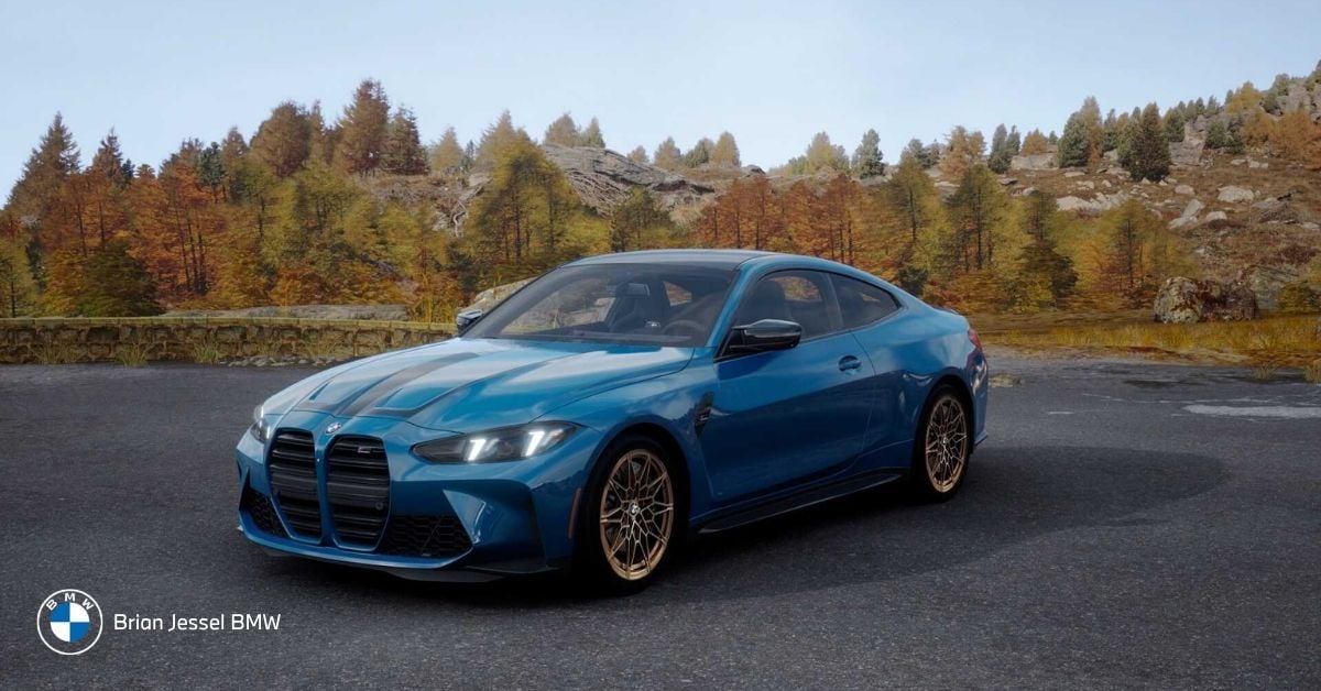 Blue BMW M4 M Legacy Edition with gold wheels parked on asphalt amid colorful autumn forest and rocky hills.