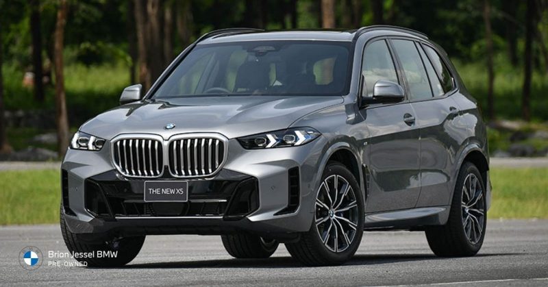 Silver BMW X5 parked on pavement with forest background, showcasing its bold front grille and athletic stance