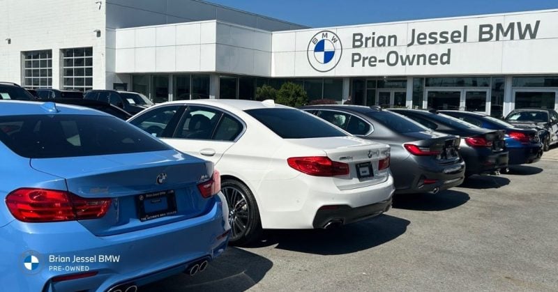 Row of BMW pre-owned cars parked in front of Brian Jessel BMW dealership building on a sunny day.