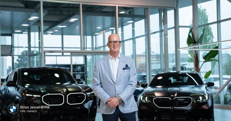 Professional standing proudly between two sleek black BMW SUVs at Brian Jessel Pre-Owned showroom in Vancouver, Canada