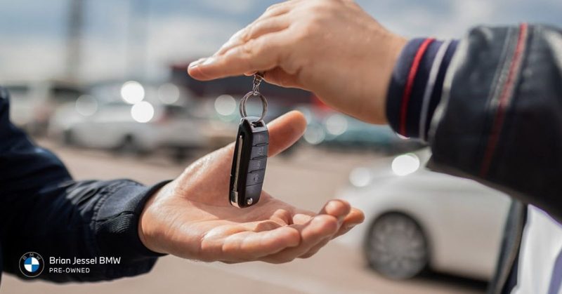 exchanging a black BMW key fob in front of a car dealership lot, highlighting pre-owned vehicle regular services