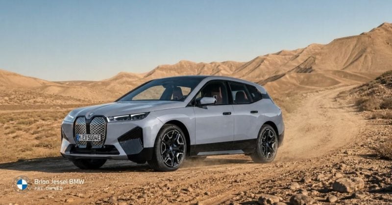 Silver BMW iX electric SUV driving across rugged desert terrain with dusty off-road conditions and mountainous background.