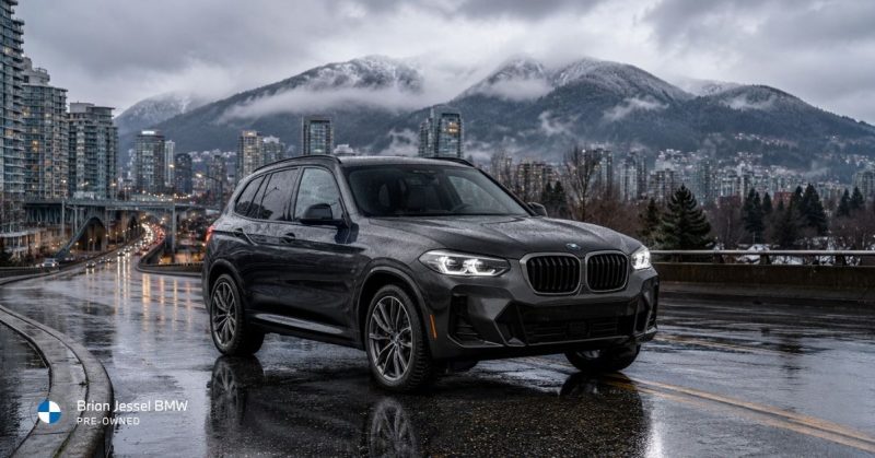 Dark gray BMW X3 xDrive SUV on a wet reflective road in Vancouver, with illuminated headlights, city skyline, bridge, and snowy mountains in the background