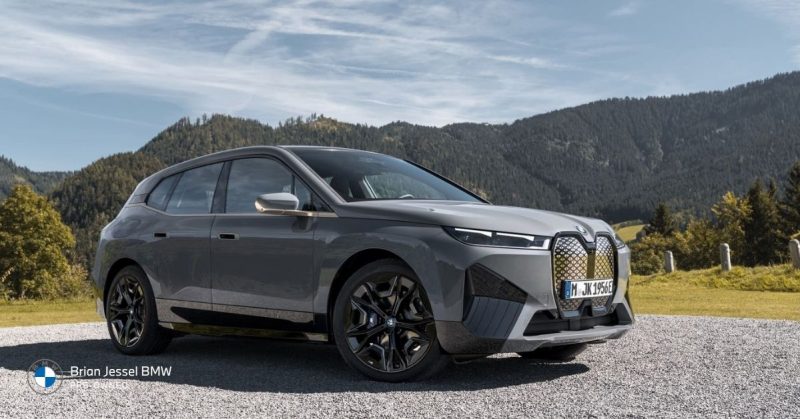 Gray BMW iX electric SUV parked on gravel with scenic mountain landscape in the background, highlighting its modern luxury design