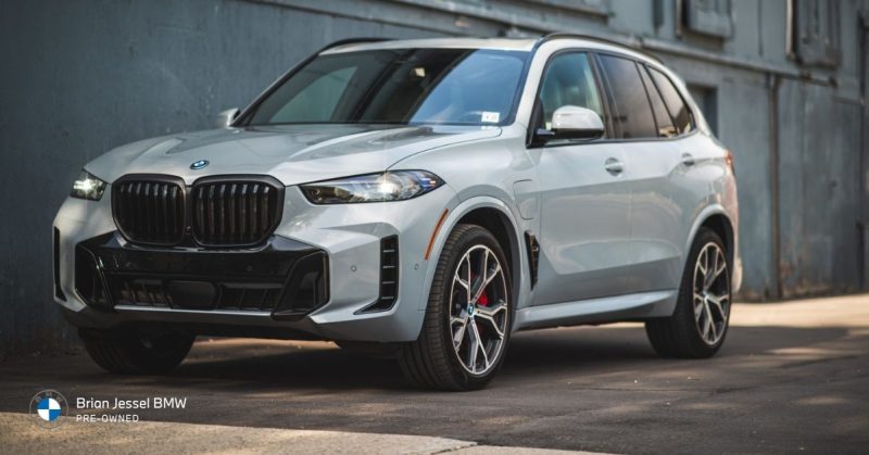 Silver BMW X5 xDrive plug-in hybrid SUV parked on pavement in front of a modern building, highlighting its sleek design and aggressive front styling