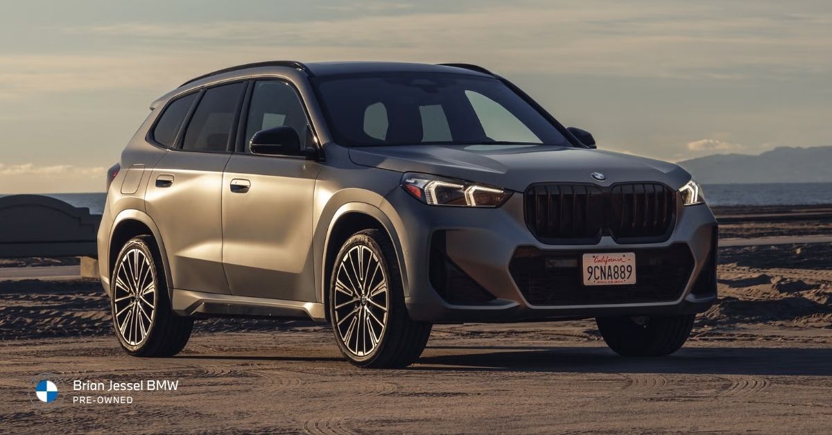 Beige BMW X1 SUV parked on a scenic beach at golden hour, with ocean and mountains in the background
