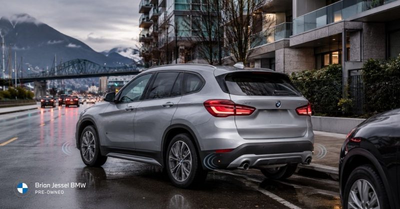 Silver BMW X4 SUV parked on a wet Vancouver street at dusk, with illuminated taillights, city buildings, bridge, and mountains in the background