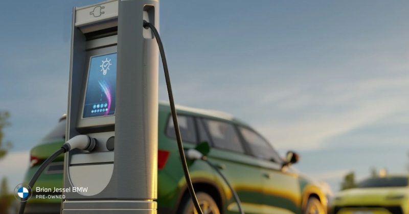 EV charging station in foreground with cable connected to a green BMW electric SUV in the background, under a clear blue sky
