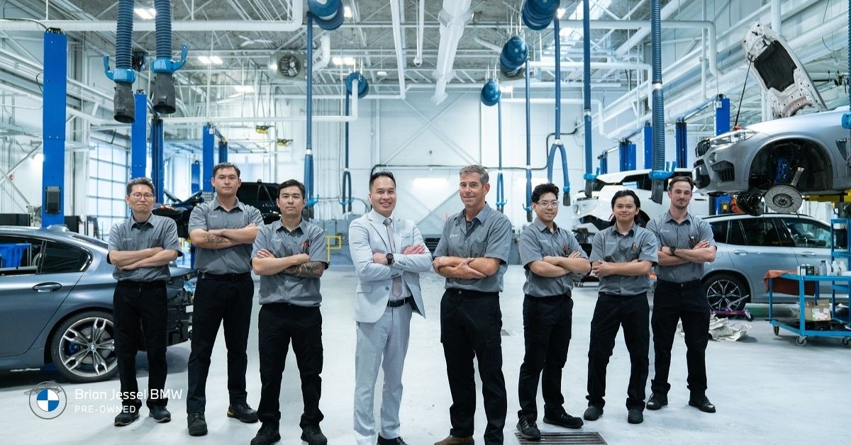 Brian Jessel BMW Pre-Owned team of sales and service staff posing together in the bright, professional service center workshop surrounded by BMW vehicles