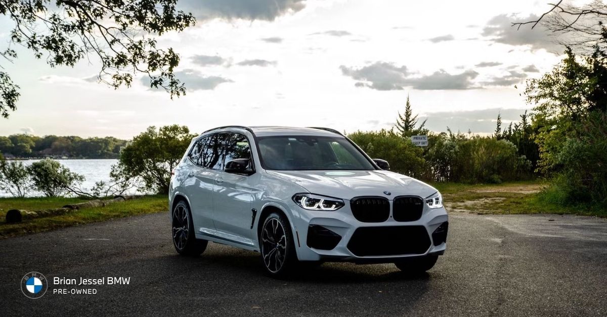 White BMW X3 SUV parked on a scenic lakeside road at dusk, surrounded by trees and waterfront with dramatic evening sky
